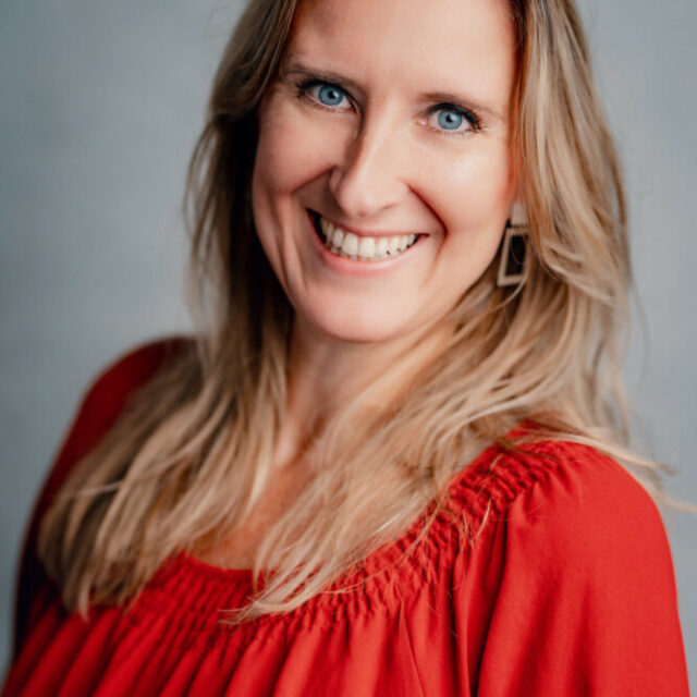 Eine Frau mit langen blonden Haaren und blauen Augen lächelt in die Kamera. Sie trägt eine rote Bluse und geometrische Ohrringe und steht vor einem schlichten grauen Hintergrund.