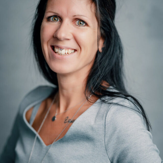 Eine Frau mit langen dunklen Haaren und hellen Augen lächelt in die Kamera. Sie trägt ein hellgraues Oberteil und eine Halskette und steht vor einem schlichten grauen Hintergrund.