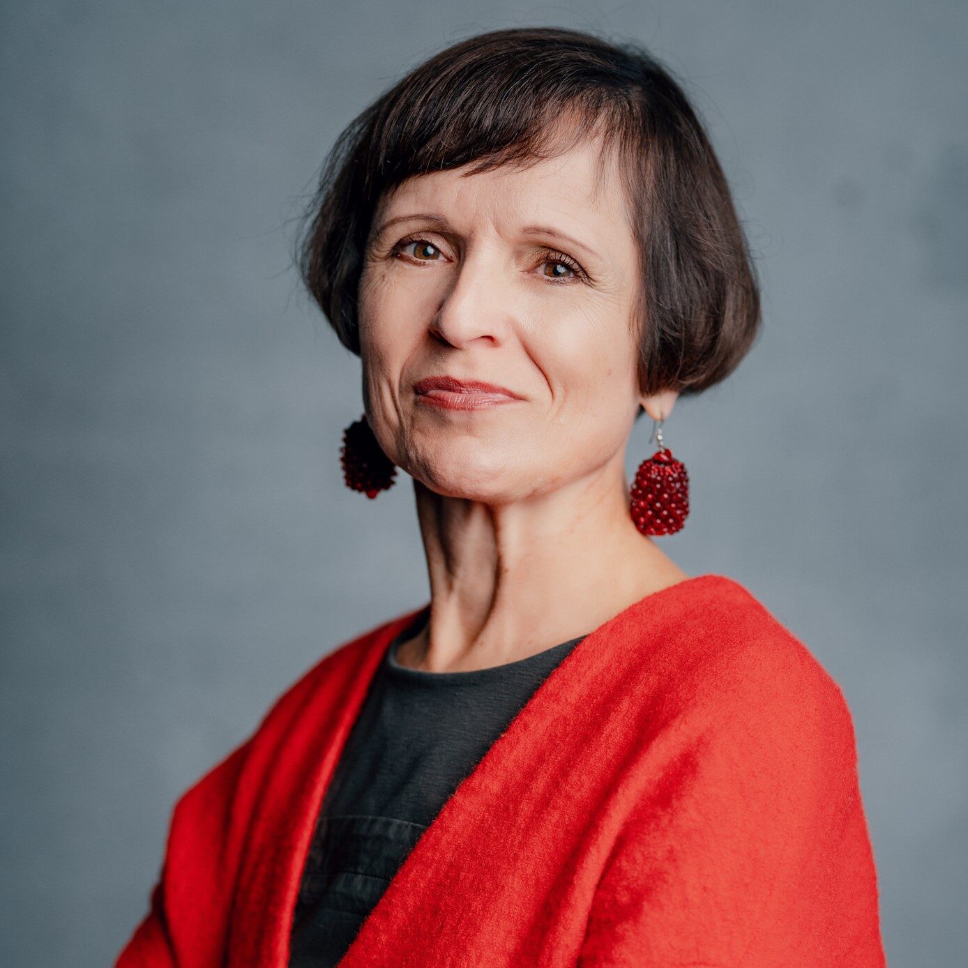 Eine Frau mit kurzen braunen Haaren, roten Ohrringen und einem leuchtend roten Pullover steht mit verschränkten Armen da und blickt selbstbewusst in die Kamera vor einem grauen Hintergrund.