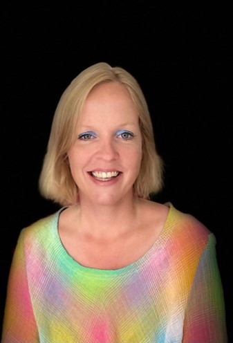 Eine lächelnde Frau mit kurzen blonden Haaren, die leuchtend blaues Augen-Make-up und ein buntes, regenbogengemustertes Oberteil trägt, steht vor einem soliden schwarzen Hintergrund.