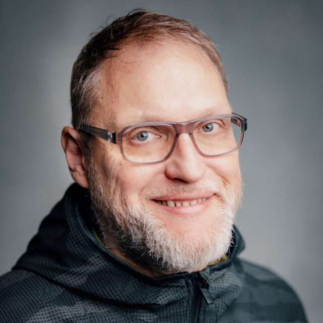 Ein Mann mittleren Alters mit kurzen hellen Haaren, einem Bart und einer Brille lächelt in die Kamera. Er trägt eine dunkle, gemusterte Adidas-Jacke mit Reißverschluss. Der Hintergrund ist sanft verschwommen und grau.