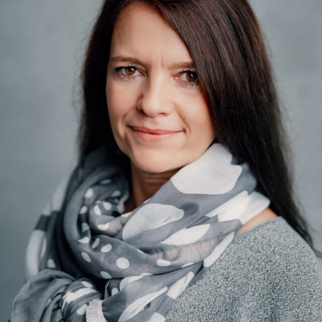 Eine Frau mit langen braunen Haaren trägt einen grauen Pullover und einen grauen Schal mit weißen Punkten und lächelt sanft in die Kamera vor einem schlichten grauen Hintergrund.