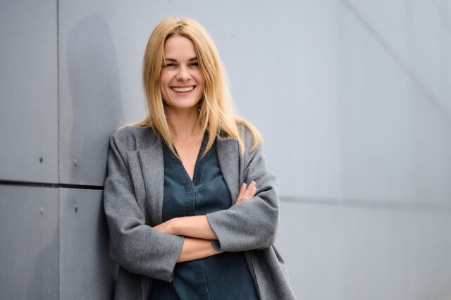 Eine Frau mit blondem Haar steht lächelnd an einer grauen Wand, die Arme verschränkt, und trägt einen grauen Mantel über einem dunklen Hemd.