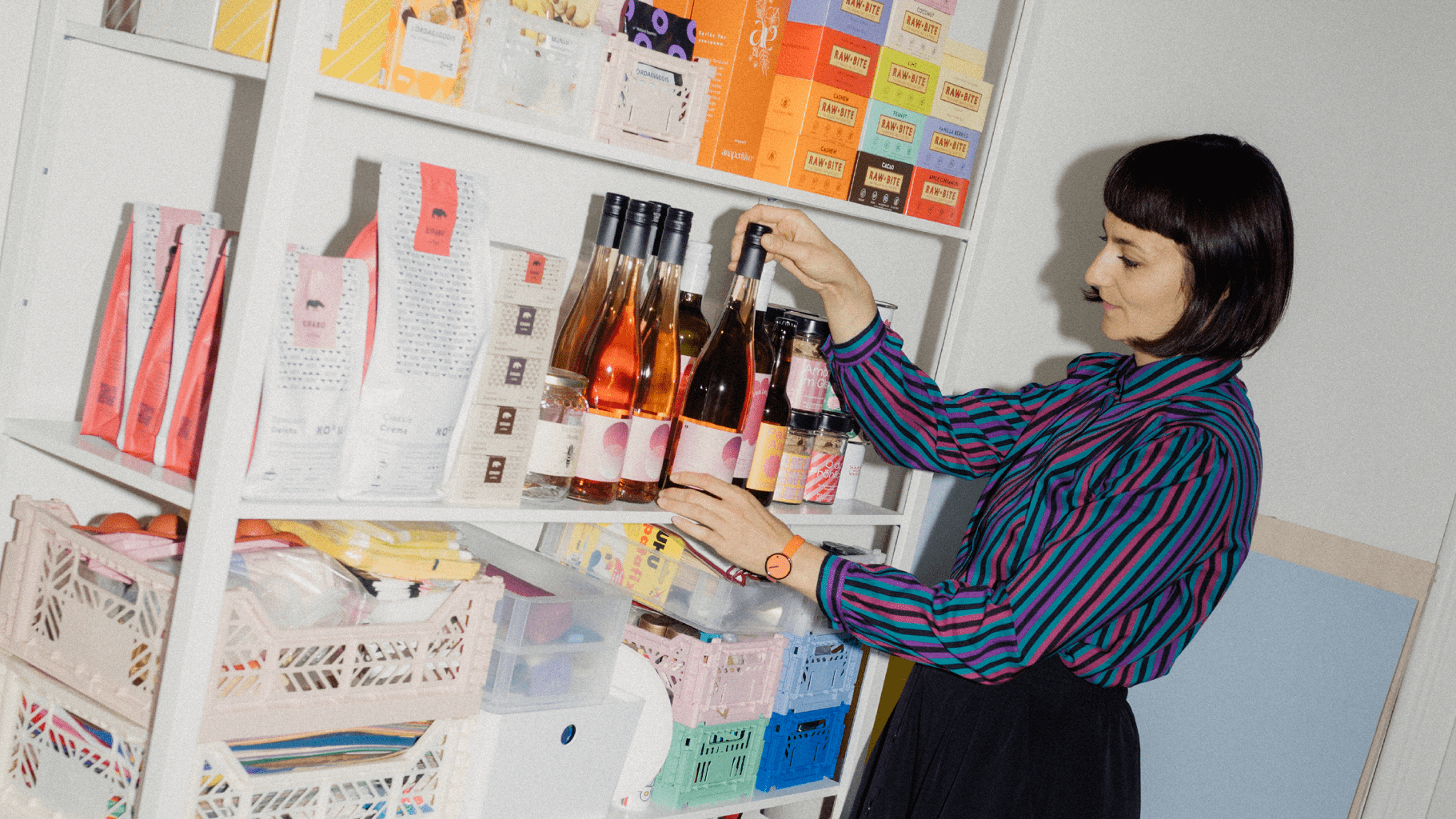 Eine Frau in einem gestreiften Hemd ordnet Weinflaschen auf einem weißen Regal, das mit verpackten Waren und Lagerkästen gefüllt ist.