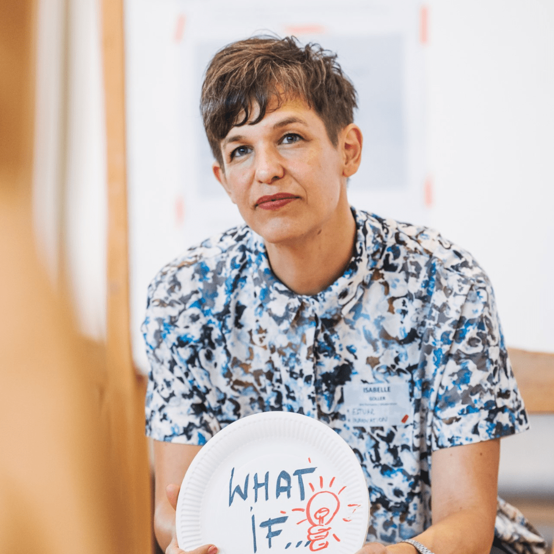 207 Eine Person mit kurzen braunen Haaren, die ein gemustertes Hemd und ein Namensschild trägt, sitzt in einem Raum und hält einen Pappteller mit der Aufschrift "WHAT IF" und einer Glühbirnenzeichnung. Sie sieht nachdenklich und engagiert aus und verkörpert den Geist von we:transform.