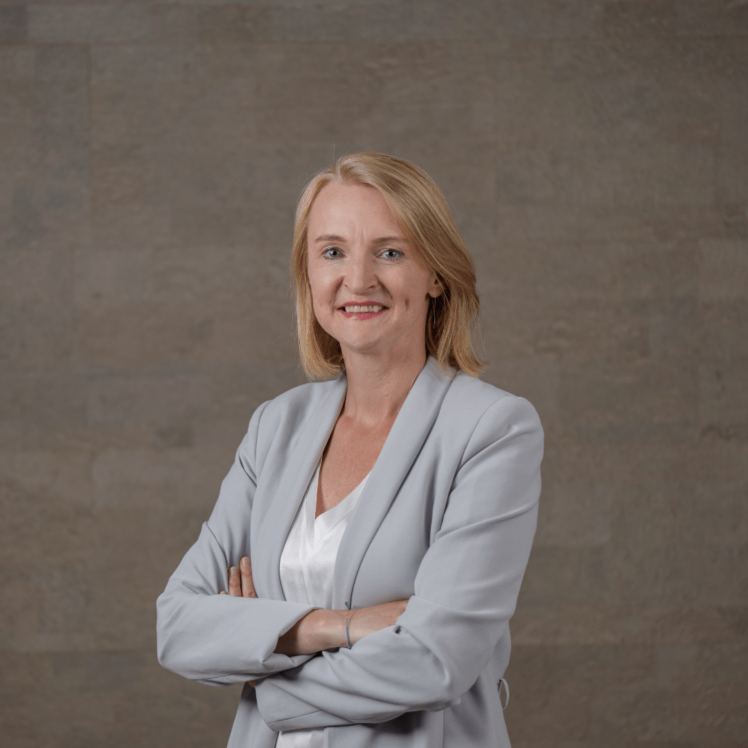 207 Eine Frau mit blondem Haar in einem hellgrauen Blazer und einer weißen Bluse steht mit verschränkten Armen und einem selbstbewussten Lächeln vor einem schlichten braunen Hintergrund und verkörpert den Geist von we:transform.