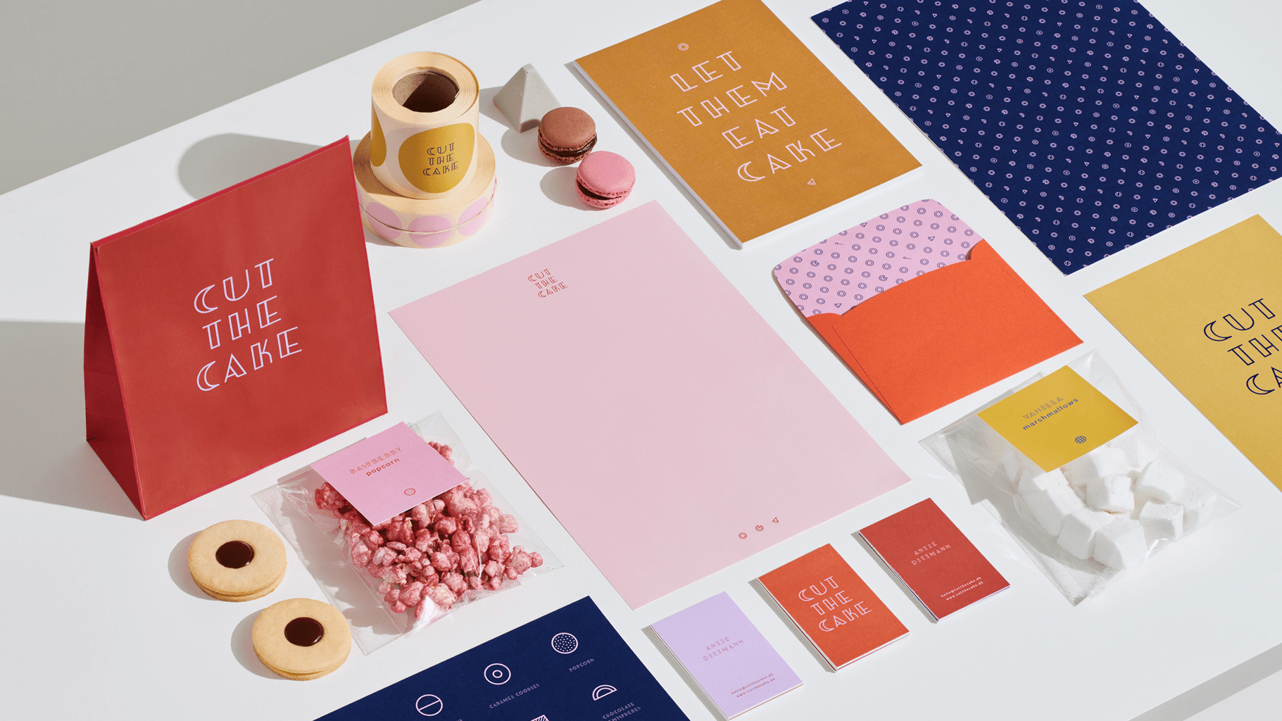 Eine flache Anordnung von farbenfrohem Markenbriefpapier und Verpackungen, einschließlich Tüten, Umschlägen, Karten, Süßigkeiten, Keksen und Macarons, die alle den Schriftzug "CUT THE CAKE" oder dekorative Muster aufweisen und ordentlich auf einer weißen Fläche angeordnet sind.