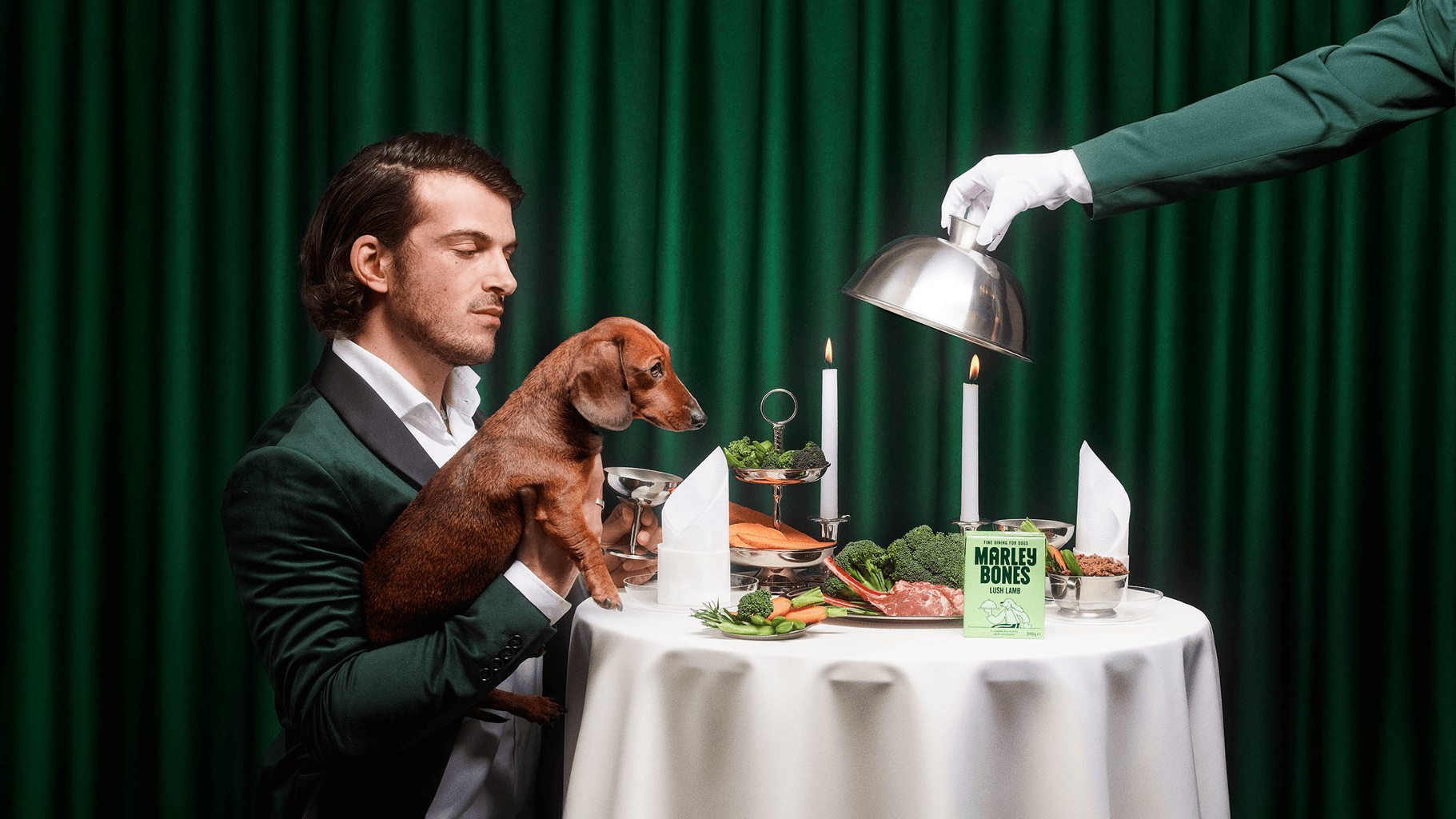 Ein Mann in einem grünen Anzug hält einen Dackel an einem schicken Tisch mit Essen, Kerzen und Servietten. Eine behandschuhte Hand hebt eine Cloche an und enthüllt ein Leckerli. Eine Schachtel mit der Aufschrift Marlis Bones steht auf dem Tisch. Im Hintergrund sind grüne Vorhänge zu sehen.