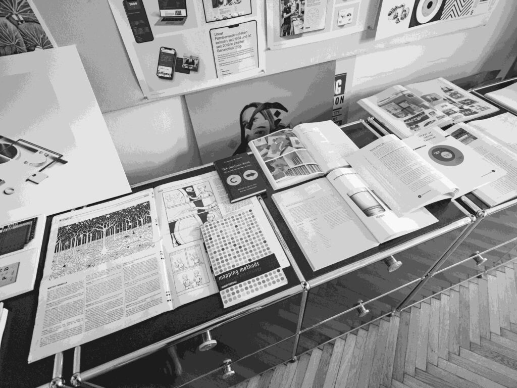 Ein monochromes Bild, das einen Ausstellungstisch mit aufgeschlagenen Büchern, Zeitschriften und Designmaterialien zeigt, auf denen Seiten mit Text, Grafiken und Illustrationen zu sehen sind. Im Hintergrund sind an der Wand befestigte Tafeln mit weiteren Materialien zu sehen.