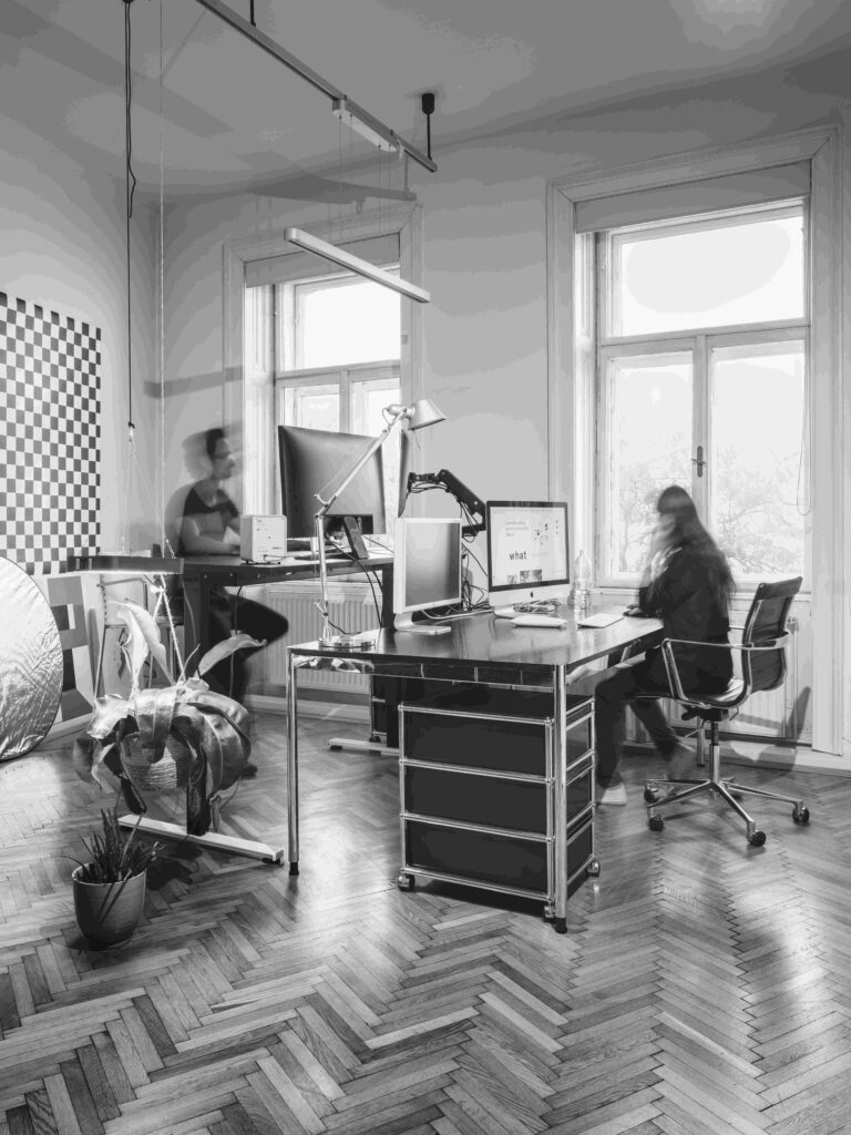 Schwarz-Weiß-Foto eines modernen Büros mit zwei Personen, die an Schreibtischen in der Nähe großer Fenster arbeiten. Eine Person sitzt an einem Computer, während die andere steht. Der Raum hat hölzerne Fischgrätenböden und eine minimalistische Einrichtung.