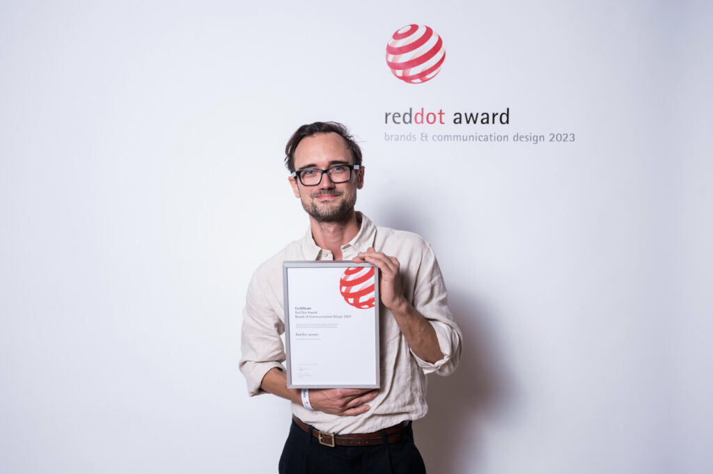 Ein Mann mit Brille und hellem Hemd hält eine gerahmte Red Dot Award-Urkunde in der Hand, während er vor einer Kulisse mit dem Red Dot Award 2023-Logo steht.