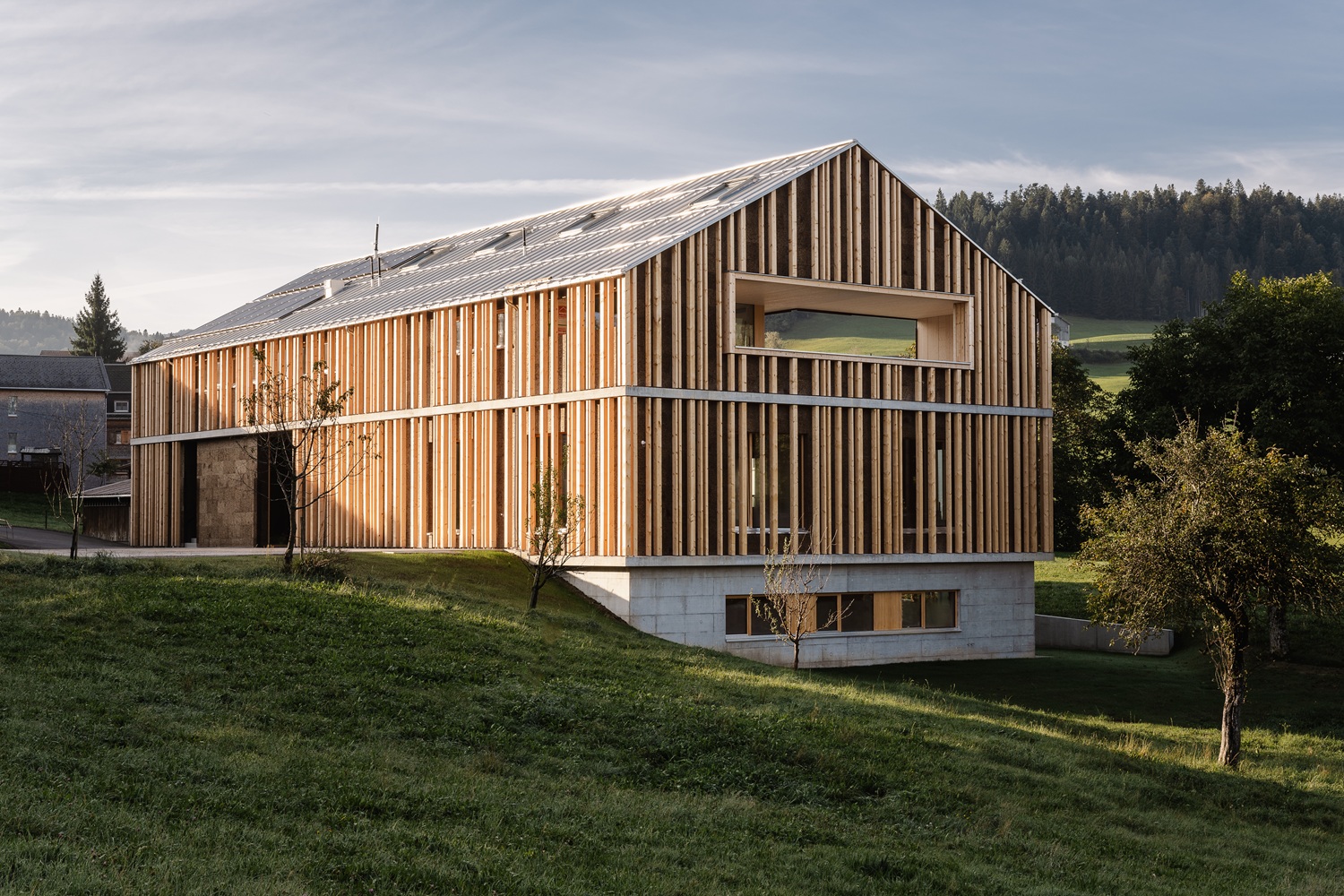 Ein modernes Gebäude im Stil einer Scheune mit vertikalen Holzlatten, großen Fenstern und einem Metalldach steht auf einem grasbewachsenen Hang - der Entwurf von Clarissa Steurer fügt sich nahtlos in die Bäume und den bewaldeten Hügel im Hintergrund ein.