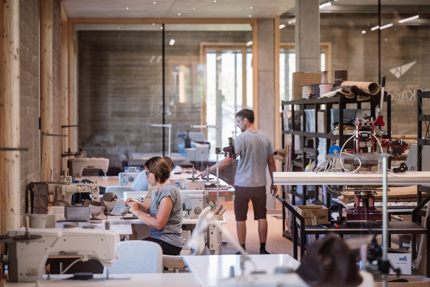 Zwei Personen arbeiten in einer hellen, modernen Werkstatt mit Holzbalken und Nähmaschinen. In den Regalen finden Materialien wie Kork Platz, und große Fenster lassen natürliches Licht in den Raum und schaffen eine handgemachte Atmosphäre mit Herz.