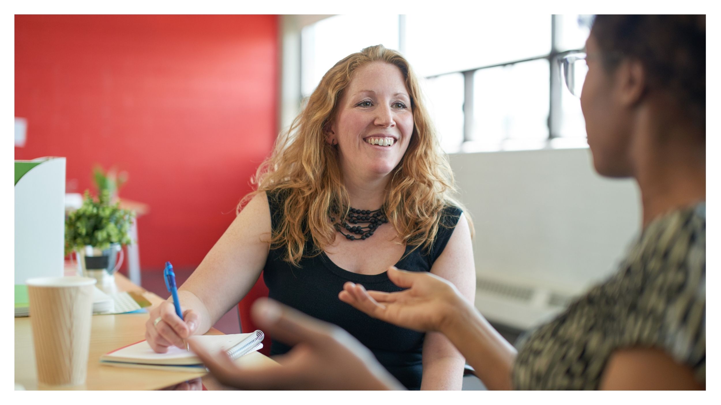 Zwei Frauen sitzen an einem Tisch und unterhalten sich. Die eine Frau lächelt, hält einen Stift und ein Notizbuch in der Hand und hört aufmerksam zu, während sie bei einem Kaffee, umgeben von Pflanzen, über die CCS-Garantiefazilität diskutieren.