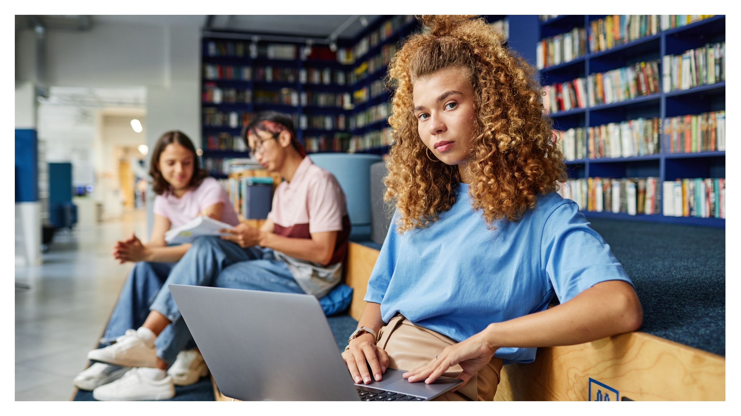 A young woman with curly hair uses a laptop in a library and looks into the camera. Two people are sitting on benches in the background, reading and talking, the walls are lined with bookshelves.