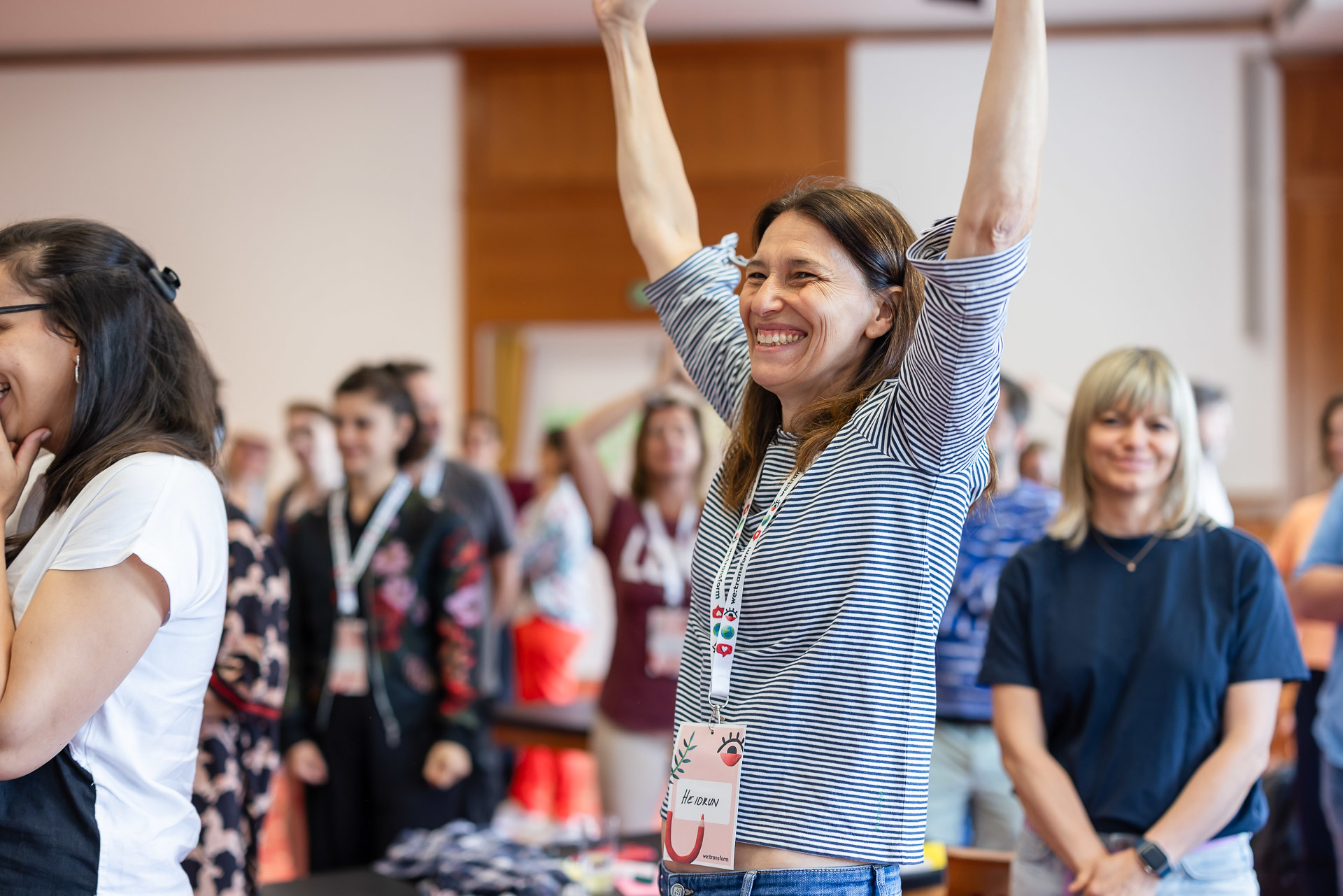 Eine Frau mit einem Namensschild steht in einem Raum voller Menschen, lächelt strahlend und hebt feierlich die Arme. Auch die anderen um sie herum wirken glücklich und engagiert. Die Umgebung sieht aus wie eine lockere Gruppenveranstaltung oder ein Workshop.