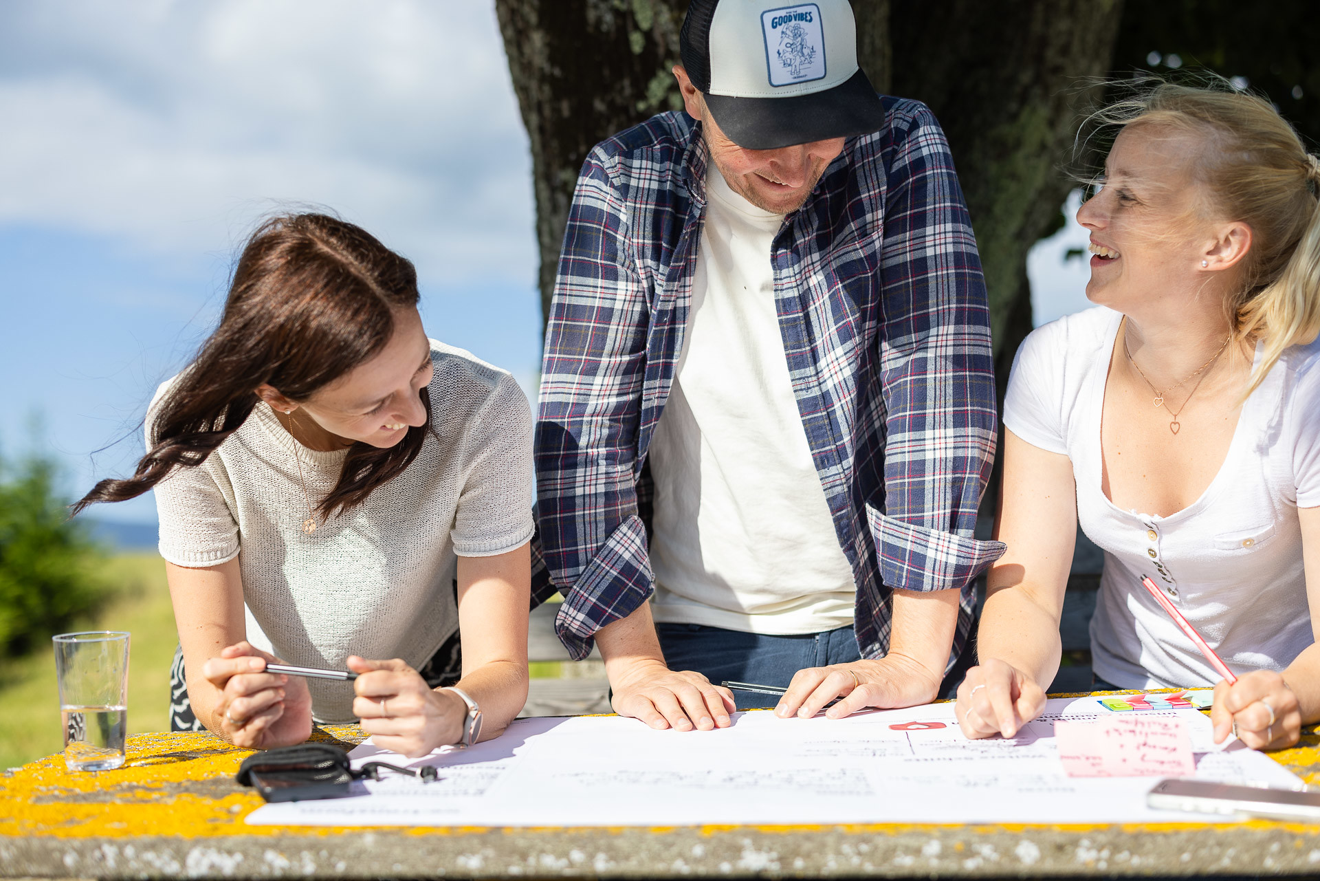 Drei Personen sitzen im Freien an einem Tisch, lächeln und unterhalten sich, während sie an Papieren und Zeichnungen arbeiten. Das Sonnenlicht fällt durch die Bäume, und es scheint, als würden sie gemeinsam an einem Projekt arbeiten oder Ideen sammeln.