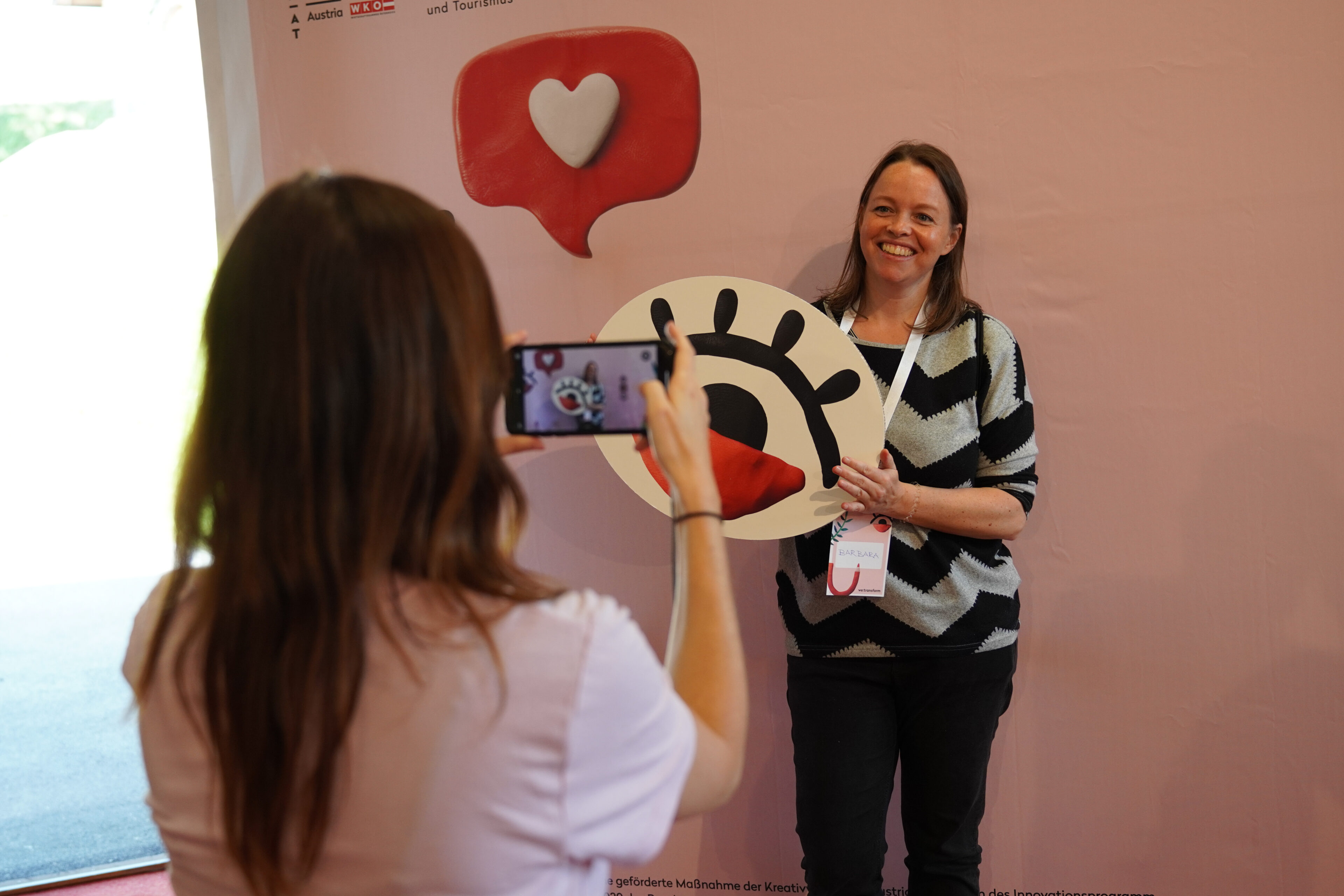Eine Frau lächelt, während sie einen großen Ausschnitt eines Auges mit herzförmiger Pupille in der Hand hält, während eine andere Person ihr Foto mit einem Smartphone vor einem rosafarbenen Hintergrund mit einem Herzsymbol macht.