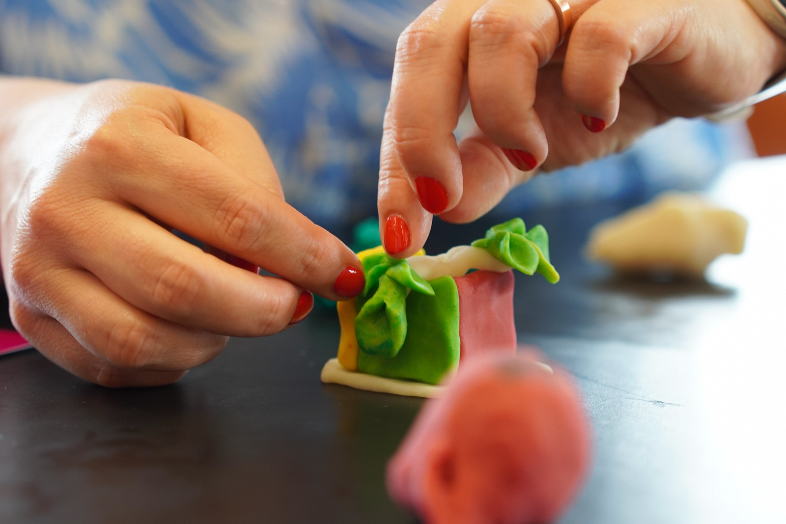 Nahaufnahme von Händen mit rotem Nagellack, die grüne Modelliermasse auf einer kleinen bunten Figur formen, mit anderen Tonformen und unscharfen Objekten auf einer dunklen Tischoberfläche.