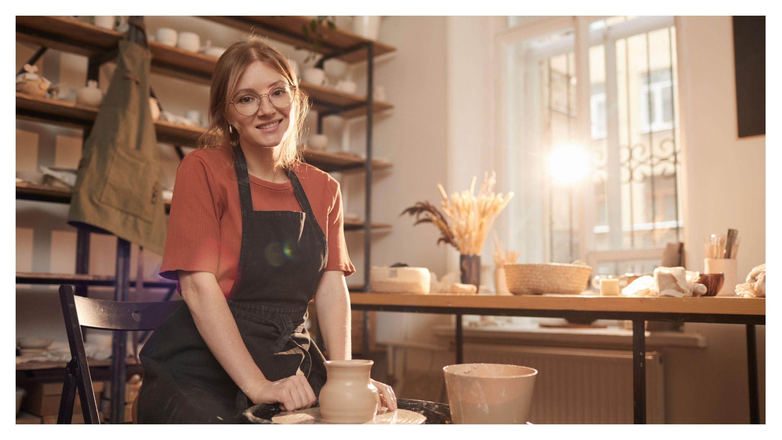 Eine Person mit Brille und Schürze sitzt lächelnd an einer Töpferscheibe in einem sonnendurchfluteten Atelier, umgeben von Keramik - ein perfekter Ort für kreative Unternehmen, um sich zu entfalten und innovative Lösungen zu entwickeln.