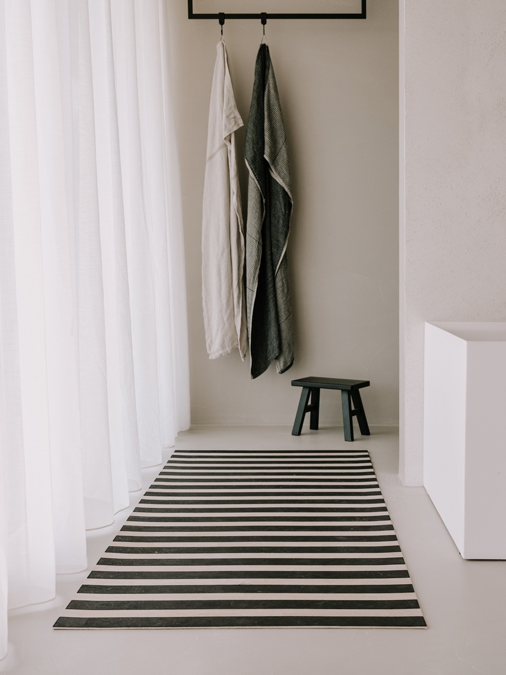 Ein minimalistisches Zimmer mit einem schwarz-weiß gestreiften Teppich von Kork, zwei Handtüchern, die an Haken hängen, einem kleinen schwarzen Hocker, durchsichtigen weißen Vorhängen und hellen Wänden und Böden.