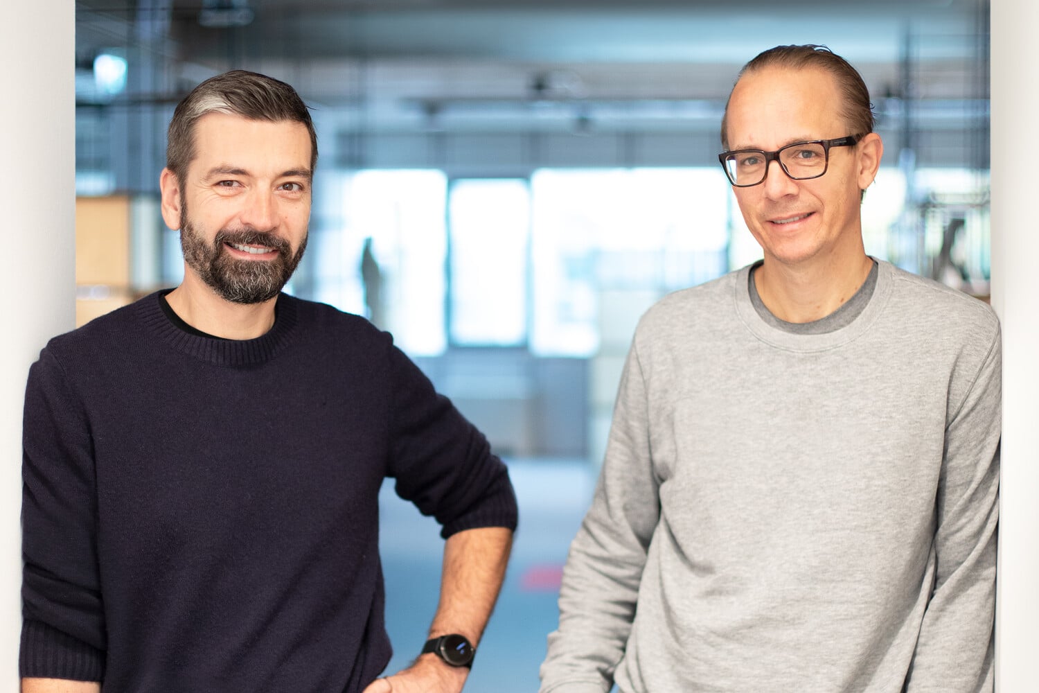 Zwei Männer stehen in einem Innenraum auf dem Creative Transformation Summit nebeneinander und lächeln in die Kamera. Der eine trägt einen dunklen Pullover und hat einen Bart, der andere eine Brille und einen hellgrauen Pullover. Der Hintergrund ist sanft verschwommen.