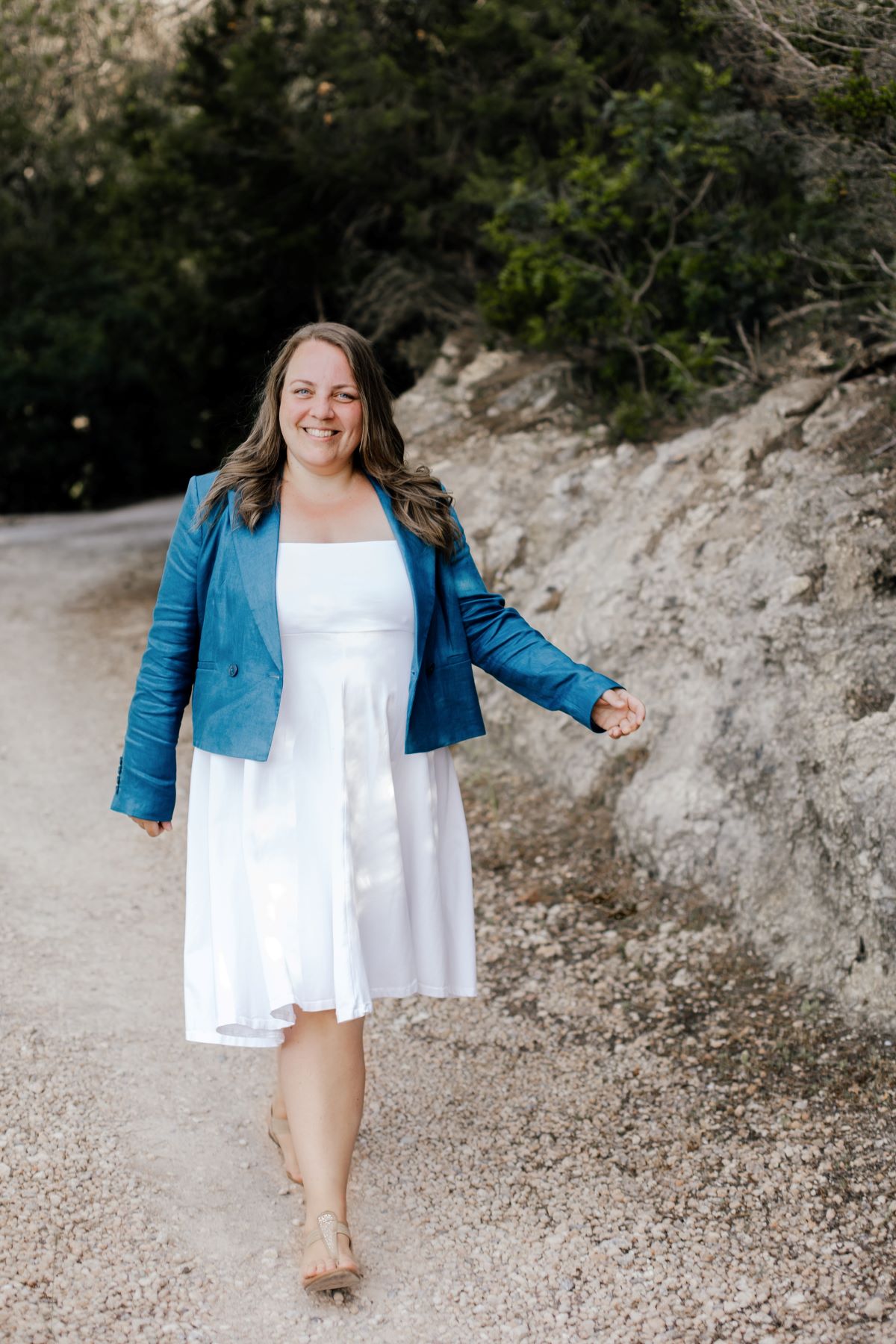 Eine Frau in einem weißen Kleid und einer blaugrünen Jacke lächelt, während sie auf einem Schotterweg neben einem felsigen Hang mit grünem Laub spaziert. Sie verkörpert den Geist von „wewant2know“, während sie ihren „Zukunftsblick“-Moment in der Natur genießt.