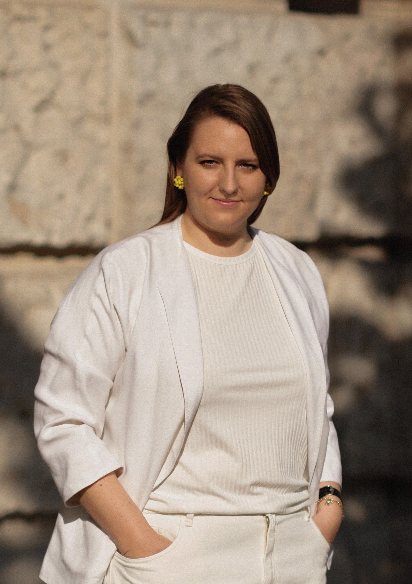 Eine Frau in weißer Jacke und weißem Oberteil steht im Freien an einer Steinmauer, während das Sonnenlicht in der Nähe Schatten wirft. Mit ihren langen braunen Haaren und den gelben Ohrringen verkörpert sie die Veränderung - mit wewant2know-Neugierde in ihrer nachdenklichen Haltung nimmt sie den Wandel an.