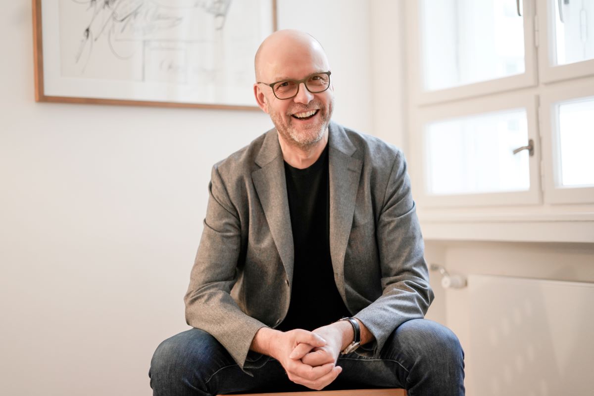 Ein lächelnder, kahlköpfiger Mann mit Brille, der einen grauen Blazer und Jeans trägt, sitzt auf einem Stuhl in einem hellen Raum mit weißen Wänden, einem Fenster und einer gerahmten Skizze im Hintergrund – ein Bild, das den Geist von „Zukunftsblick“ einfängt, während er zuversichtlich in die Zukunft blickt.