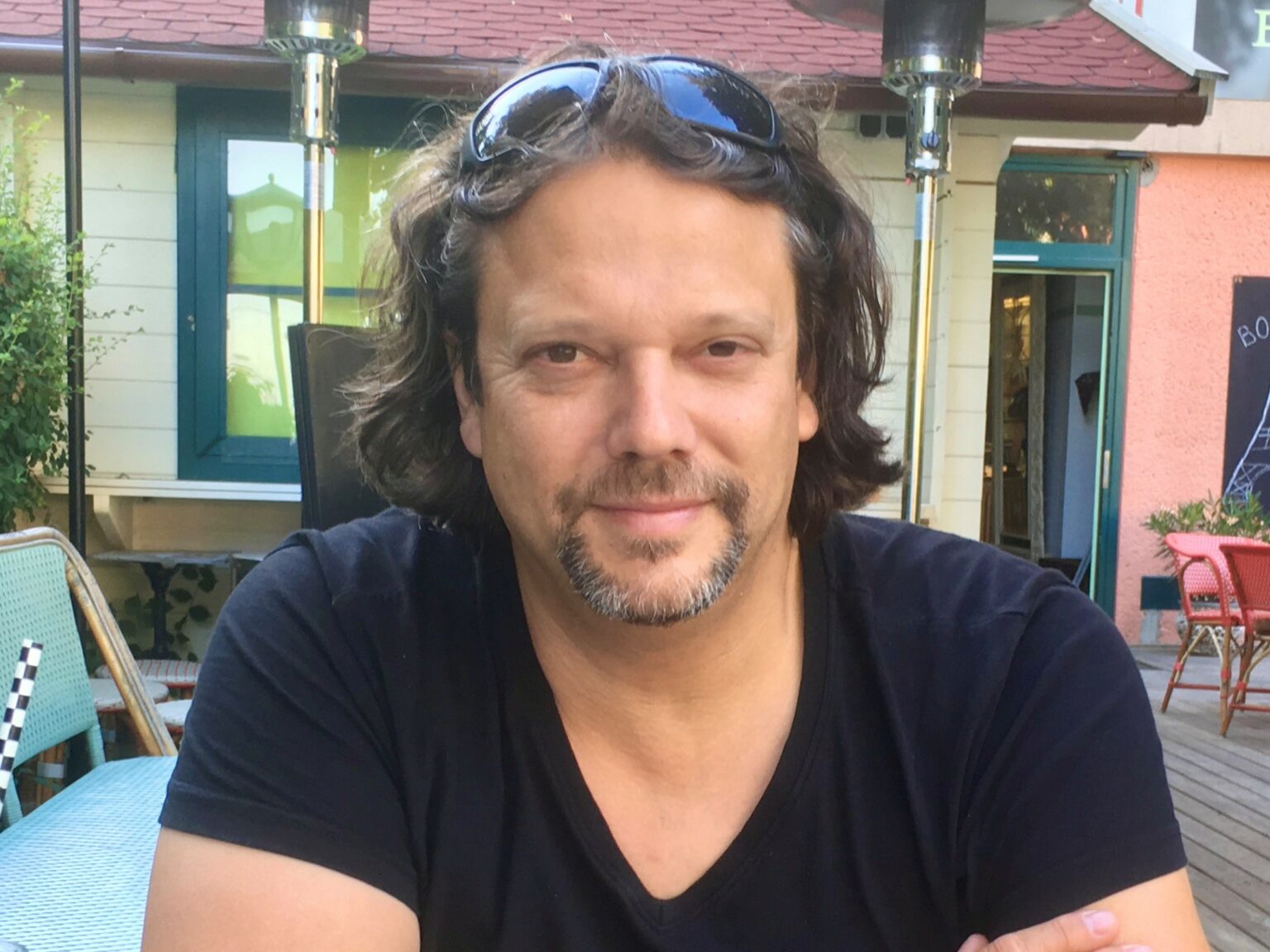 Ein Mann mit schulterlangem Haar, einer Sonnenbrille auf dem Kopf und einem schwarzen T-Shirt sitzt im Freien an einem Tisch und lächelt leicht. Im Hintergrund sind ein Gebäude und Terrassenmöbel zu sehen - ein entspannter Moment von Christian Kroepfl aus Die Zukunft ist Rund.