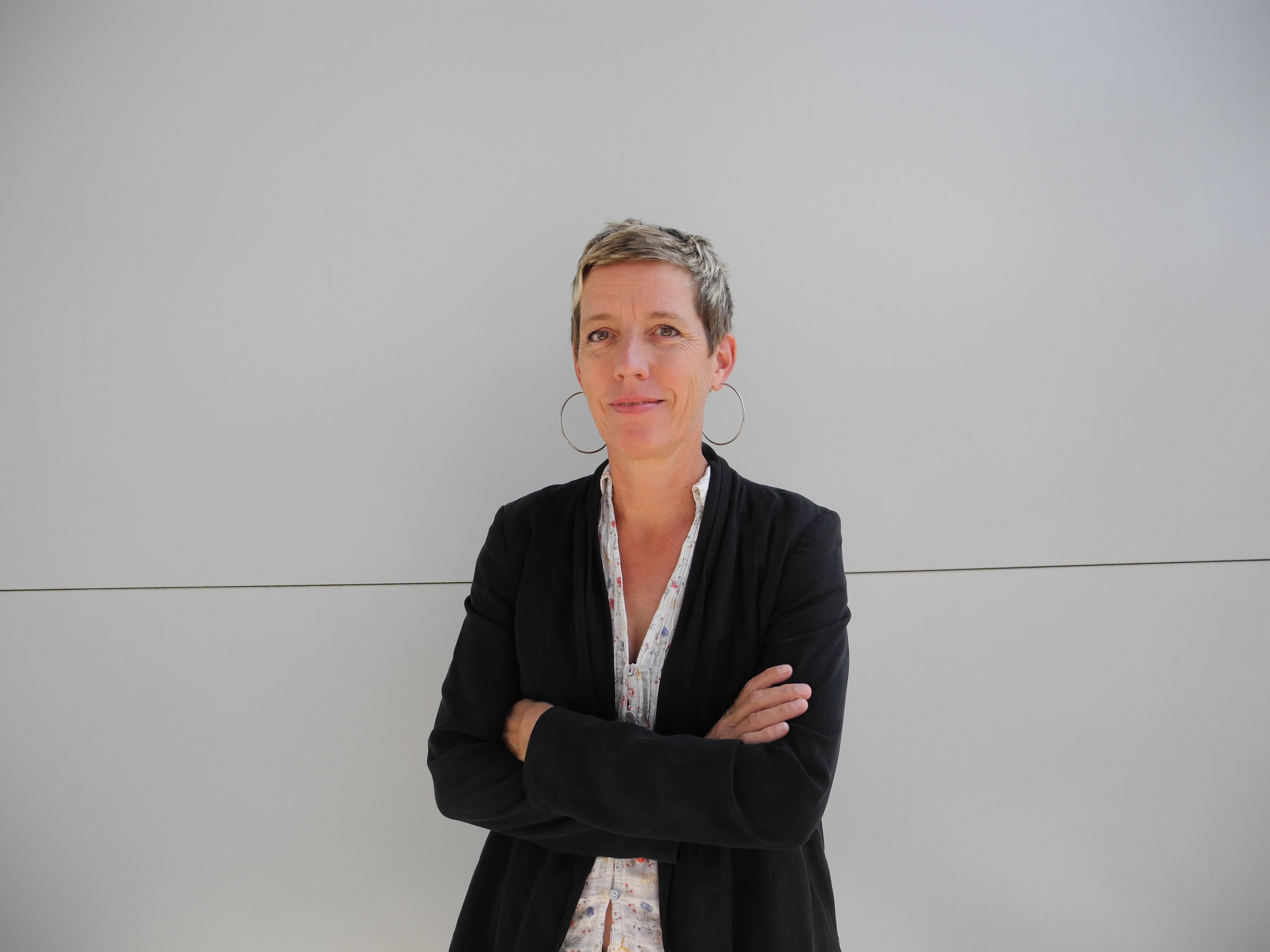 A businessman with short hair, a black blazer and earrings stands with his arms folded in front of a plain light gray wall and looks calmly into the camera.