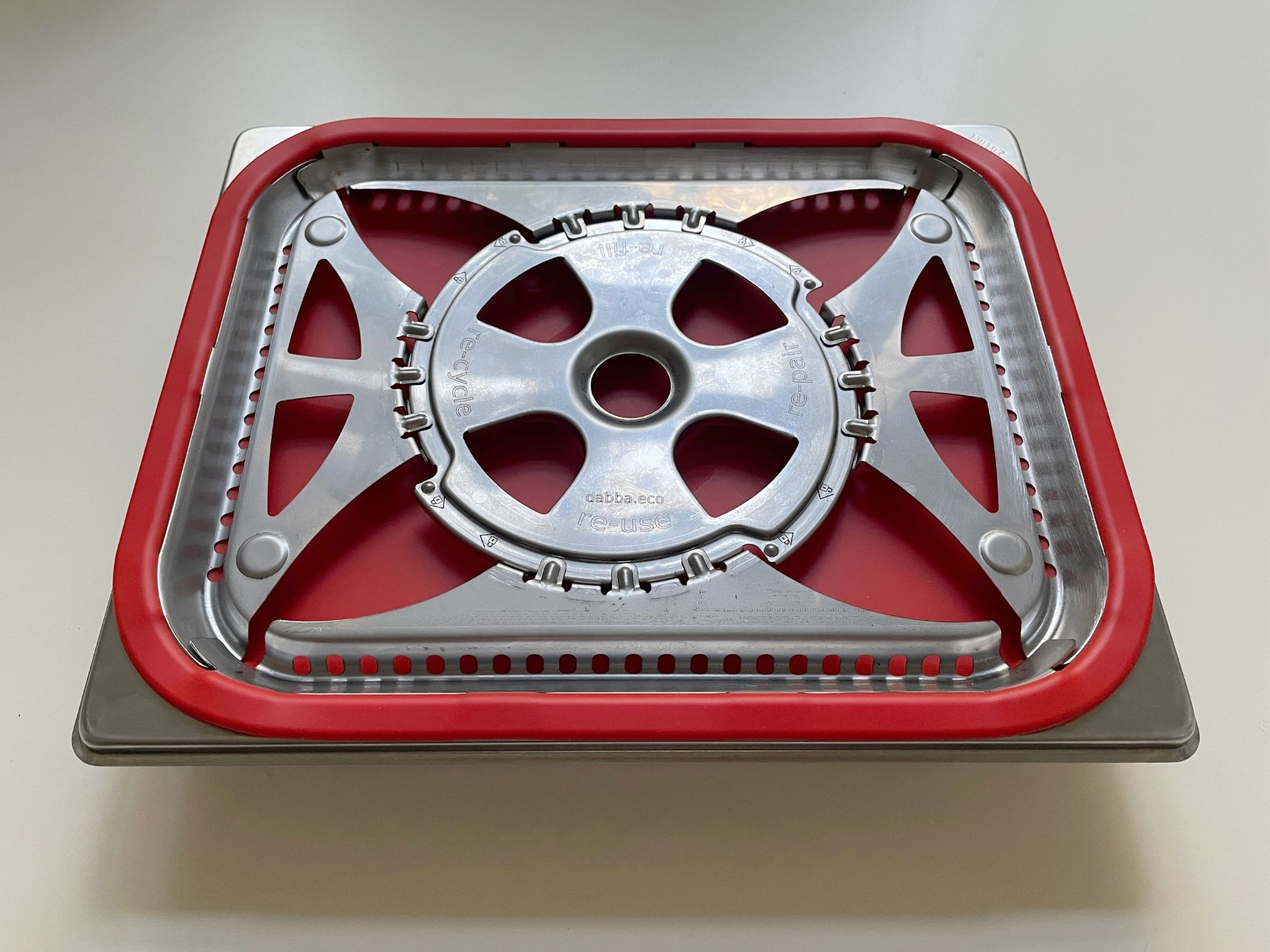 Ein Metalltablett mit rotem Silikonrand und einem runden Metallteil mit Speichen liegt darauf. Das Tablett wird auf eine helle Fläche gestellt.