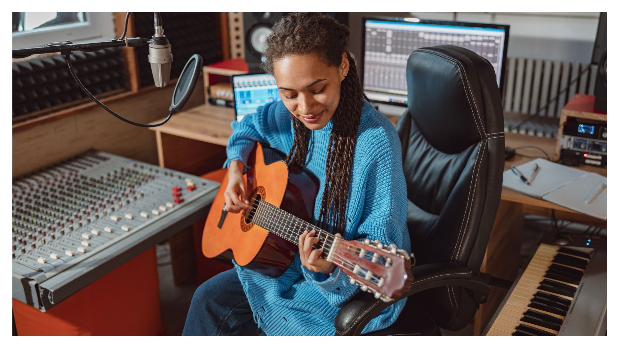 A person with long braided hair plays an acoustic guitar in a recording studio, surrounded by mixing desks and computer monitors displaying audio software - perhaps preparing for a scholarship application or a commencement fellowship.