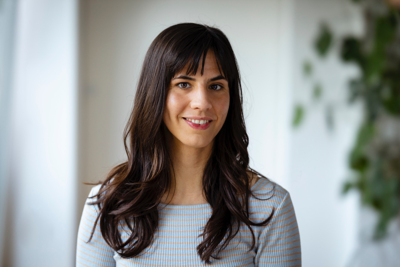 A woman with long dark hair and bangs, wearing a bright, striped top, smiles at the camera in a dimly lit interior with blurred green plants in the background.