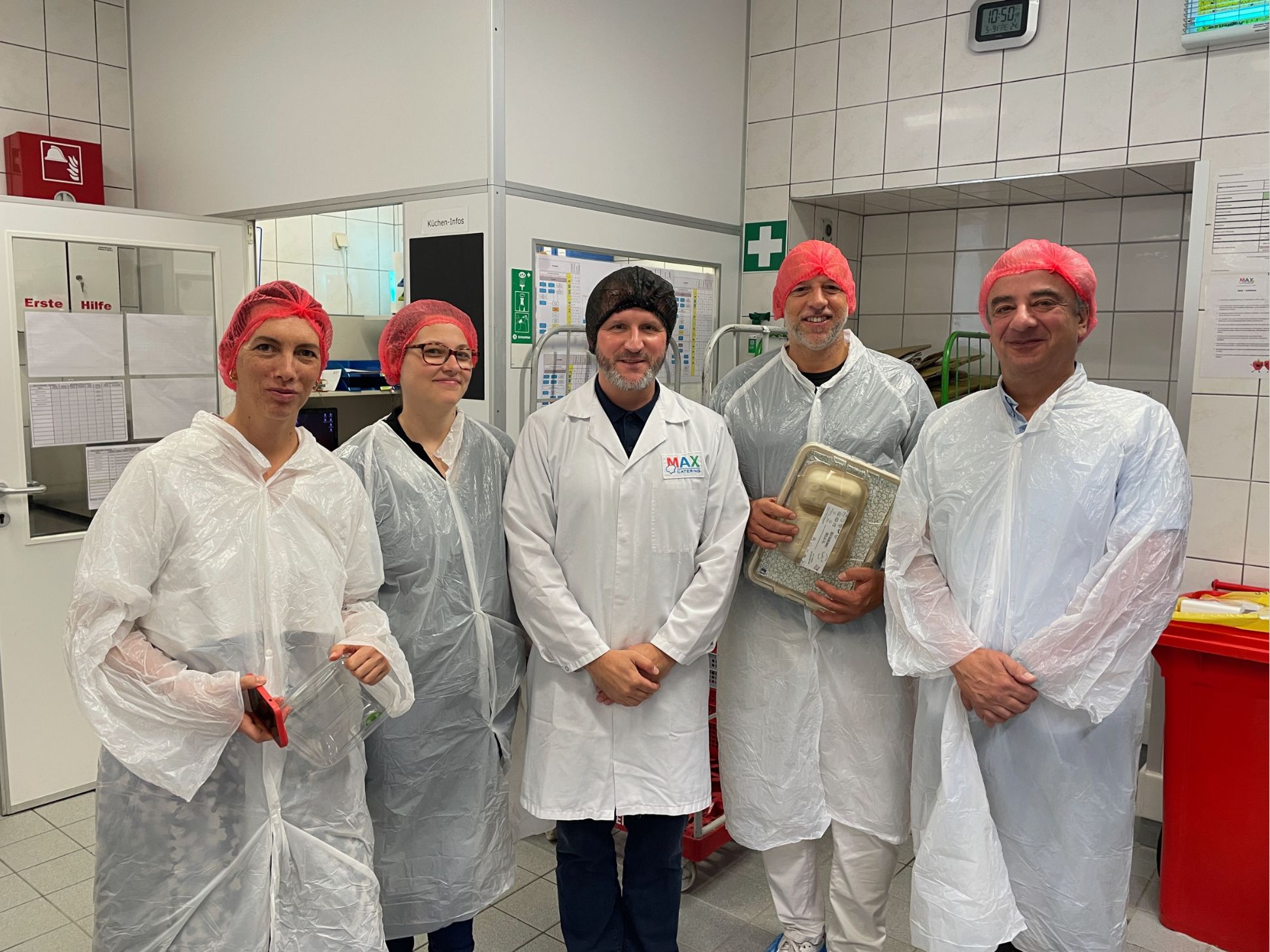 Fünf Personen in weißen Laborkitteln, Haarnetzen und Schuhüberzügen stehen in einem gekachelten Laborraum. Einer hält ein Lebensmittelpaket. Im Hintergrund sind Laborgeräte und Sicherheitsschilder zu sehen.