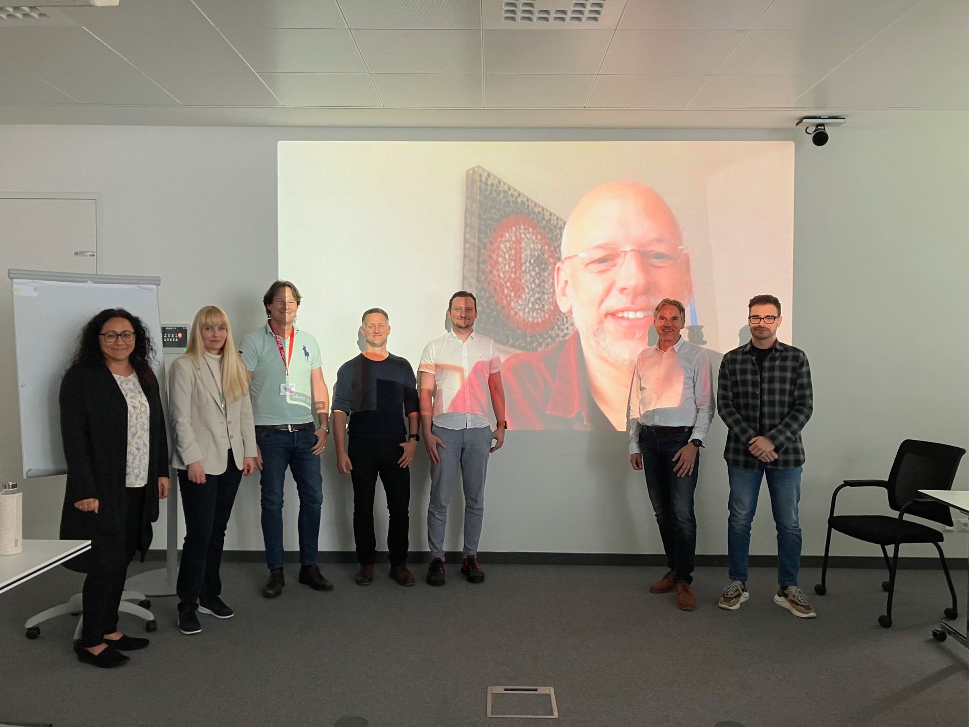 Sieben Personen stehen in einem Konferenzraum und posieren für ein Foto, während eine weitere Person per Videoanruf auf einem großen Bildschirm hinter ihnen erscheint. Im Raum befinden sich Stühle, ein Flipchart und Bürogeräte.