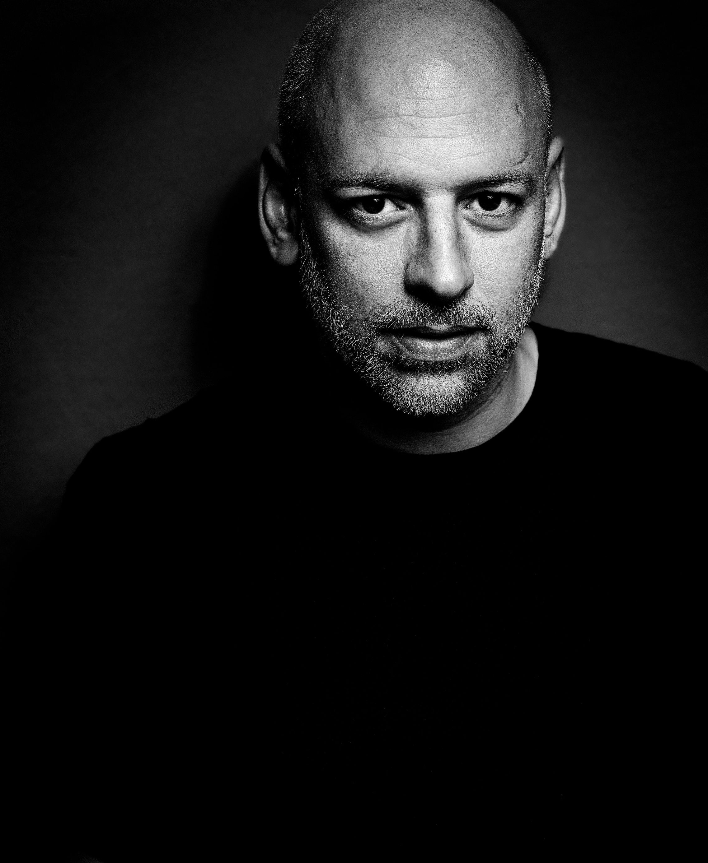 Ein Schwarz-Weiß-Porträt eines Mannes mit Glatze und Bart, der ein dunkles Hemd trägt und mit ernster Miene direkt in die Kamera schaut. Die Beleuchtung hebt eine Seite seines Gesichts hervor und erzeugt starke Schatten.