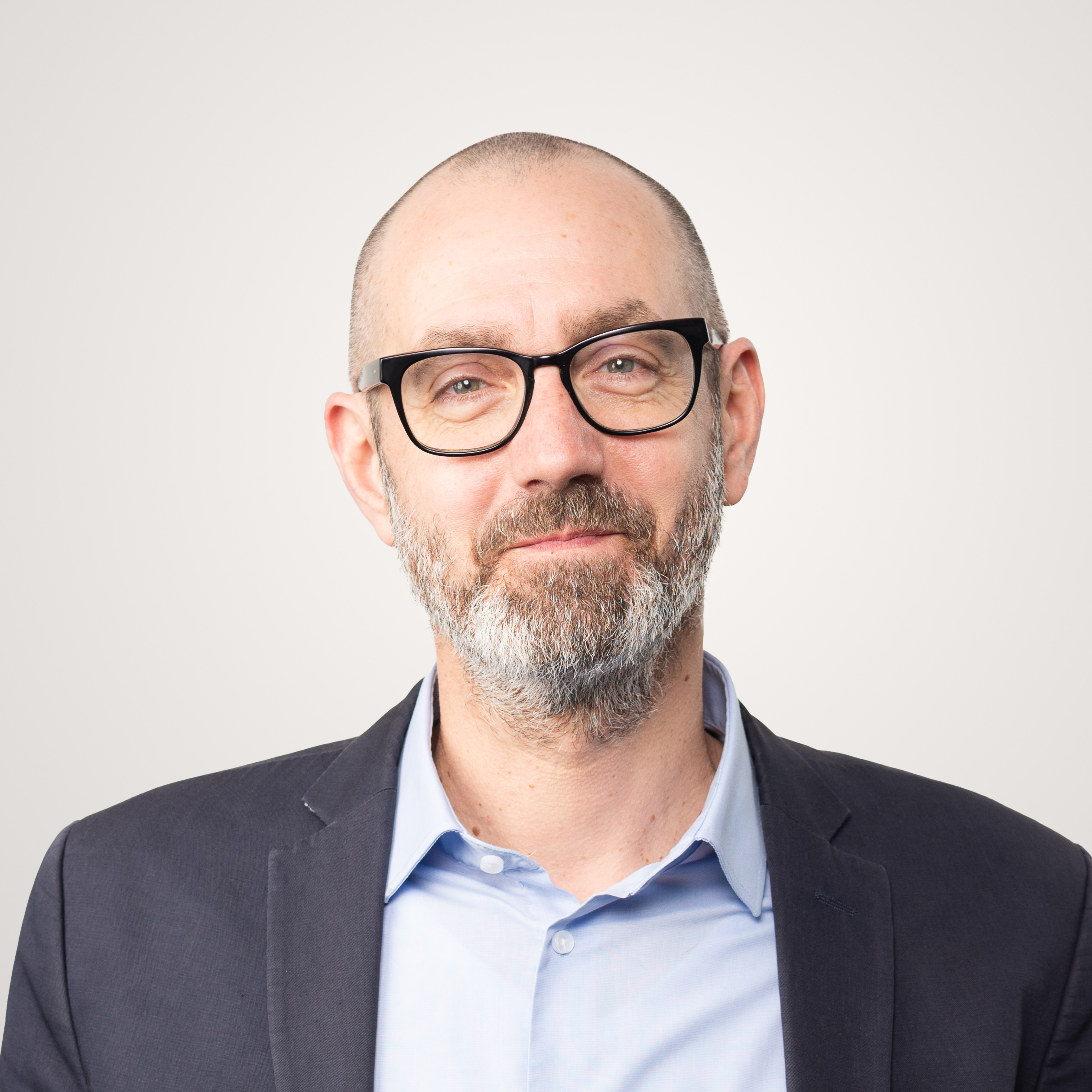 A middle-aged businessman with a bald head, short gray beard and black glasses, wearing a dark blazer over a light blue shirt, stands in front of a plain light background, as if taking a break from running in circles.