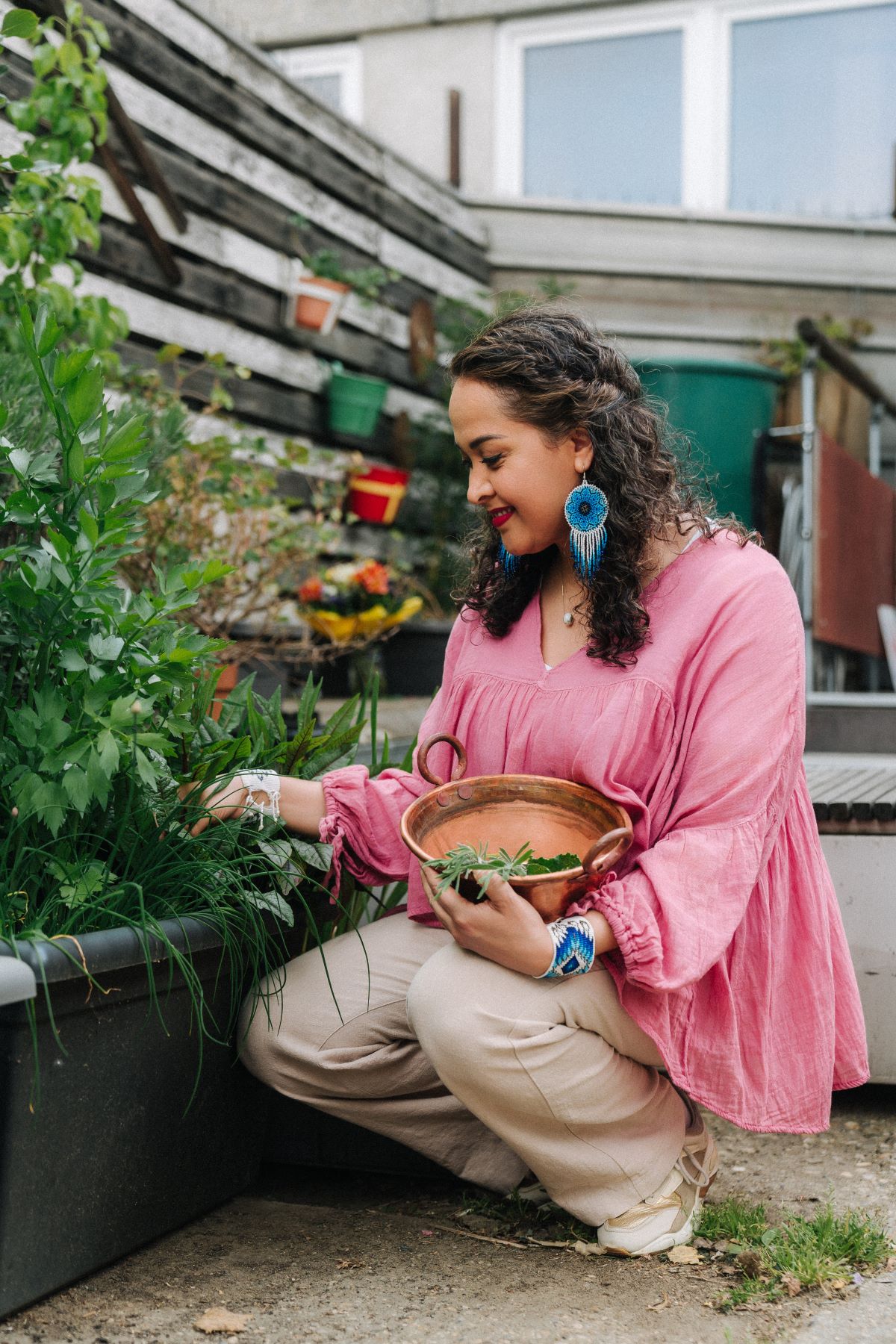 A woman in a pink blouse and beige trousers kneels by a raised bed and smiles as she picks herbs for her business and places them in a copper bowl. She is surrounded by green plants in an outdoor garden, not walking in circles but concentrating on her harvest.