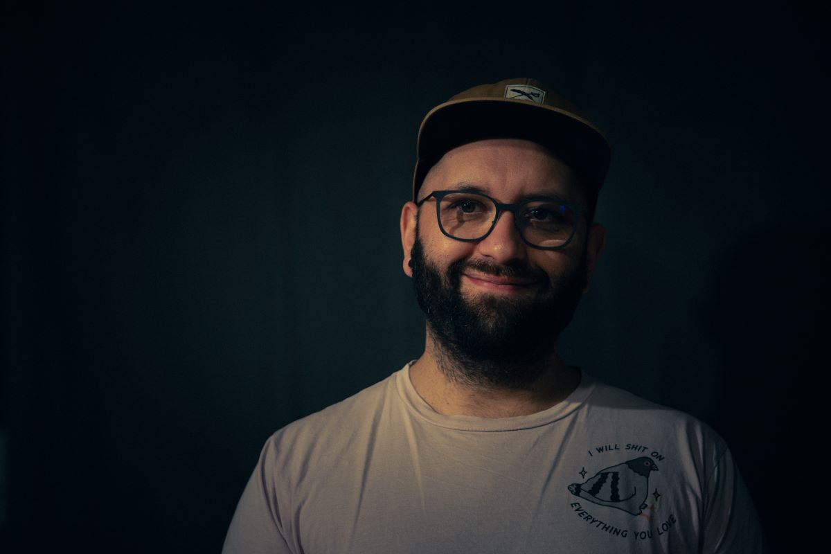 Vor einem dunklen Hintergrund steht ein lächelnder Mann mit Bart, Brille und Mütze; er trägt ein helles Hemd mit einem kleinen Motiv und dem Schriftzug „Zukunftsblick“ auf der Brust.