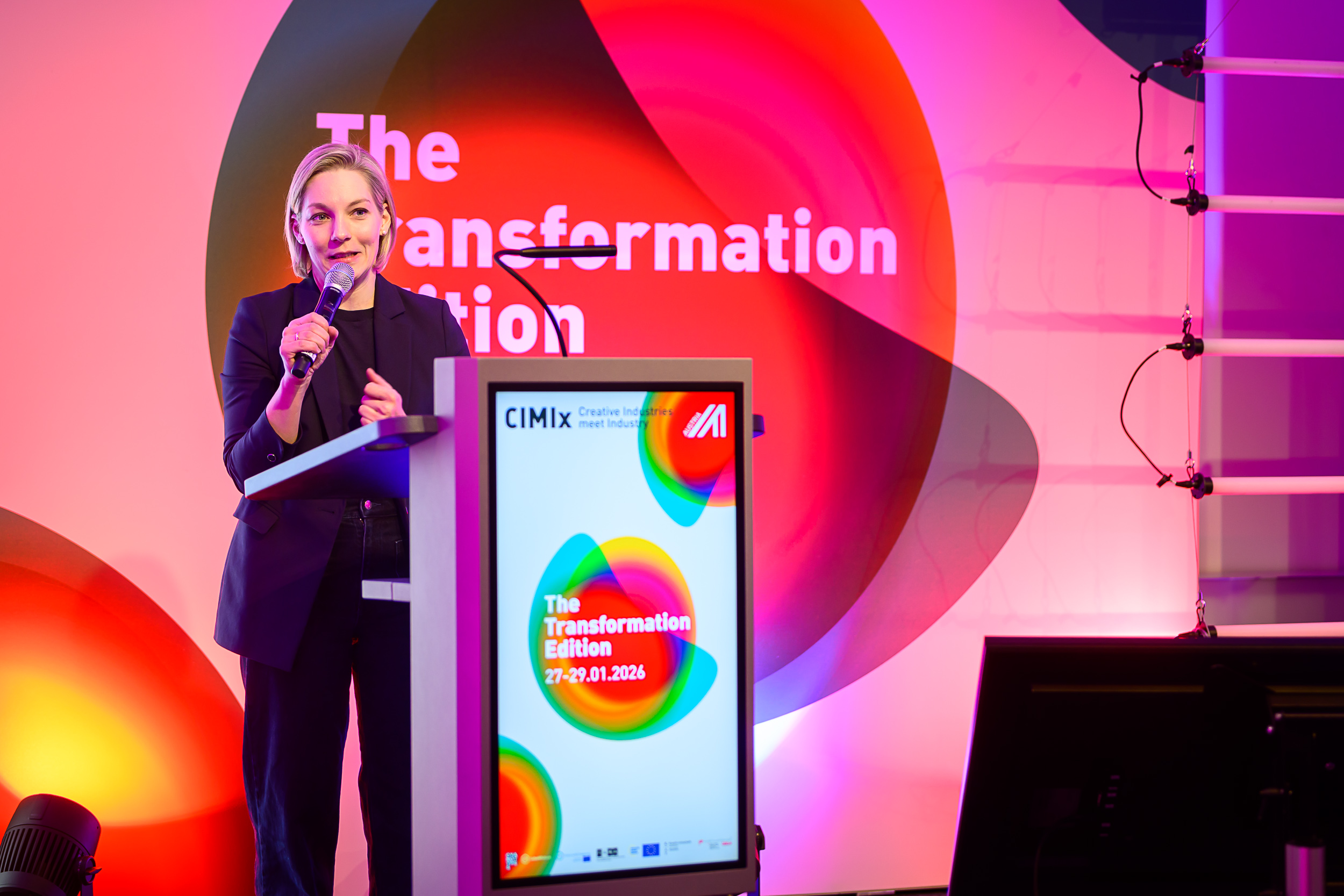 Eine Frau spricht auf einem Podium während der CIMIx 2026: The Transformation Edition in ein Mikrofon. Helle, bunte Grafiken und Veranstaltungsdetails werden auf einem Bildschirm und einem Hintergrund hinter ihr angezeigt.