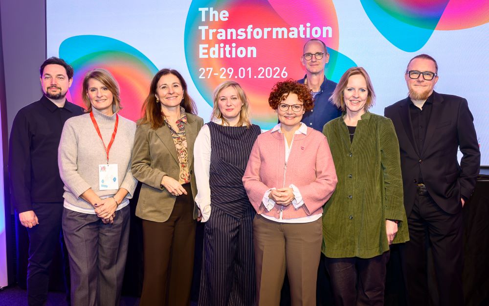 Eine Gruppe von acht Personen steht lächelnd vor einem farbenfrohen Bildschirm, auf dem "The Transformation Edition 27-29.01.2026" beim CIMIx 2026 angezeigt wird. Der Bildschirm zeigt abstrakte Formen und leuchtende Farben im Hintergrund.