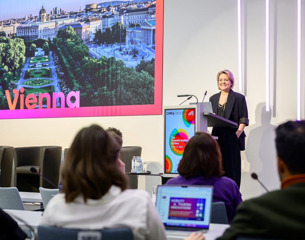 Eine Frau steht an einem Podium und hält einen Vortrag vor dem Publikum des CIMIx 2026. Ein großer Bildschirm hinter ihr zeigt ein Bild von Wien und einen Teil des Wortes Wien. Die Menschen sitzen und hören zu, einige mit geöffneten Laptops.