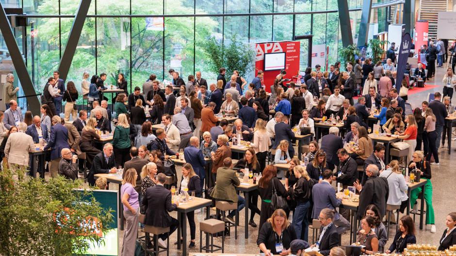 Eine gro&szlig;e Gruppe von Menschen versammelt sich in einem hellen, ger&auml;umigen Innenraum beim Forward Festival und kn&uuml;pft an hohen Tischen Kontakte. Viele sitzen oder stehen und unterhalten sich, w&auml;hrend im Hintergrund Gr&uuml;nfl&auml;chen und gro&szlig;e Fenster zu sehen sind.