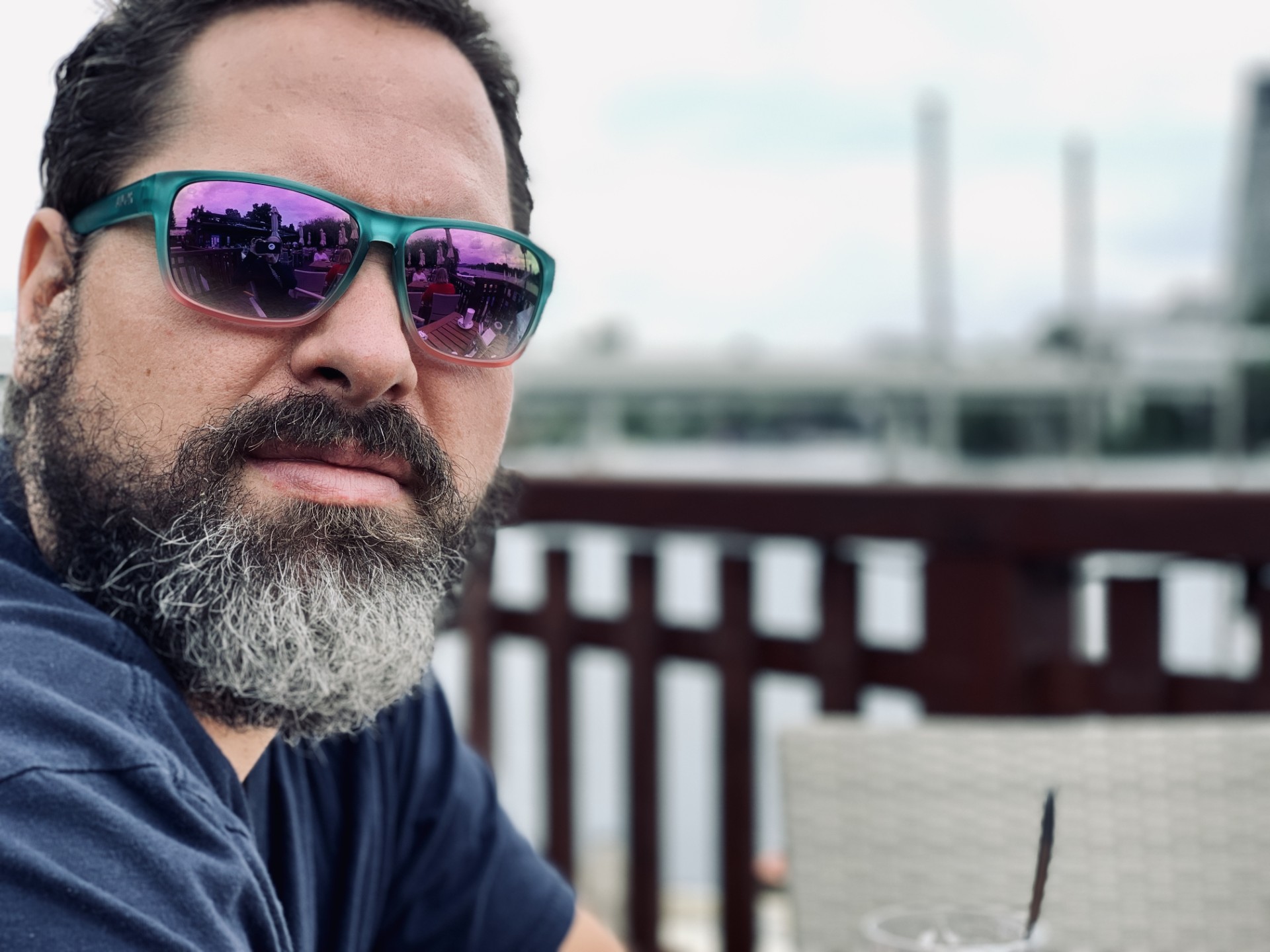 Ein b&auml;rtiger Mann mit Sonnenbrille sitzt im Freien an einem h&ouml;lzernen Gel&auml;nder, w&auml;hrend Wasser und industrielle Strukturen im Hintergrund verschwimmen. Die Reflexion in seiner Sonnenbrille zeigt Patrick Beranek, den Fotografen, und die umgebende Szene.