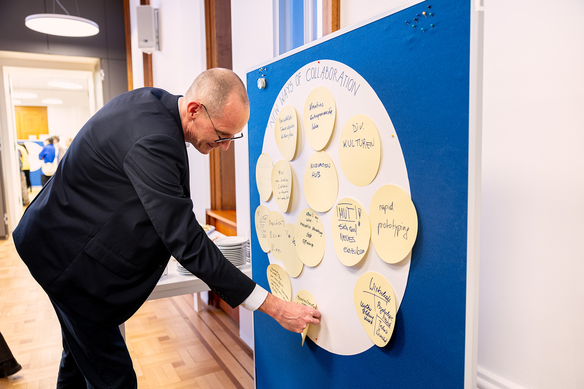 Ein Mann im Anzug legt einen gelben Klebezettel auf eine blaue Tafel, auf der kreisförmig handgeschriebene Ideen stehen. In einem hellen Raum mit Holzfußboden wird der Brainstorming-Prozess für den Kreativwirtschaftsbericht 2026 dargestellt.