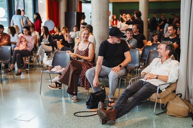 Eine Gruppe von Menschen sitzt w&auml;hrend der Vienna Design Week in einem hellen Veranstaltungsraum und entspannt sich. Drei Erwachsene in der ersten Reihe plaudern und l&auml;cheln, ein Mann l&uuml;mmelt mit ausgestreckten Beinen, w&auml;hrend sich andere im Hintergrund unter die Leute mischen.