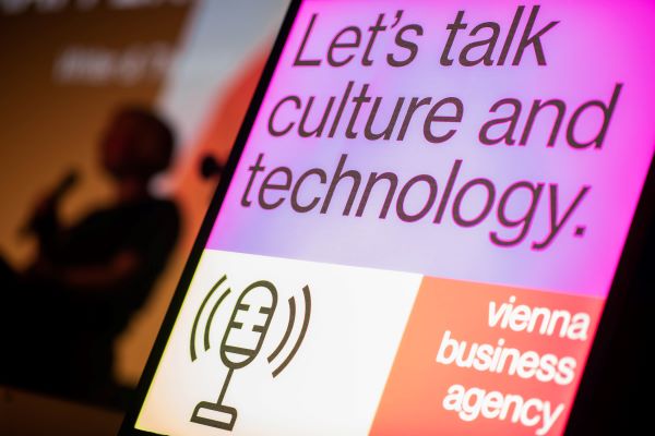 Ein Schild mit der Aufschrift "Let's talk culture and technology" (Lasst uns &uuml;ber Kultur und Technologie sprechen), ein Mikrofon-Symbol und das Logo der Wirtschaftsagentur Wien. Im Hintergrund steht ein unscharfer Redner auf der B&uuml;hne, der den globalen Dialog im Vorfeld der SXSW London 2026 hervorhebt.