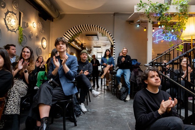 Eine Gruppe von Menschen sitzt während der Vienna Design Week in einem Innenraum auf schwarzen Stühlen, klatscht und blickt nach vorne. An der Wand hängen dekorative Teller, Pflanzen und eine Leuchtreklame mit der Aufschrift Büro. Einige der Teilnehmer sehen engagiert und aufmerksam aus.