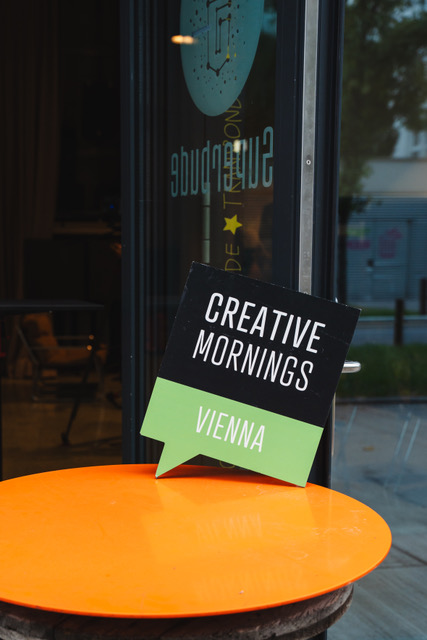 Ein schwarz-grünes Schild mit der Aufschrift Creative Mornings Vienna steht auf einem orangefarbenen Tisch vor einer Glastür und fängt den lebendigen Geist der Vienna Design Week ein.